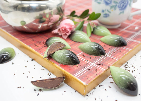 Teardrop-shaped dark chocolate bonbons with green and black coloring and a ganache filling, on a white table with teapot and pink rose