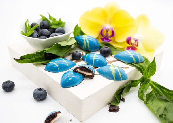 Blueberry Cream teardrop shaped dark chocolate bonbon with blue coloring and green stripes, filled with blueberry and white chocolate ganache on a white table with blueberries and basil leaves