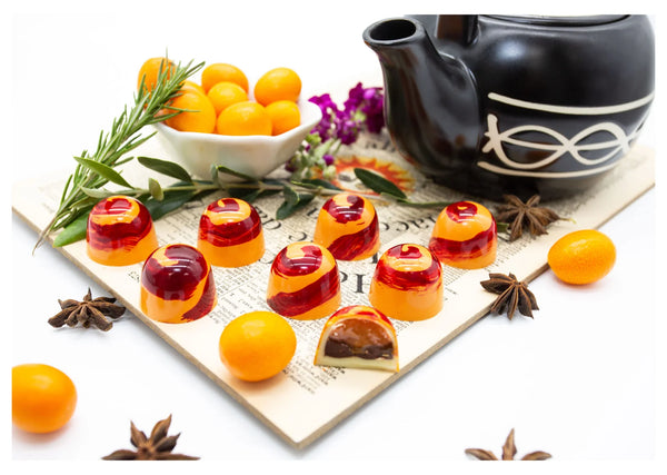 Gumdrop-shaped chocolates with a red and orange spiral design, filled with kumquat marmalade and ganache on a white table next to star anise and kumquat