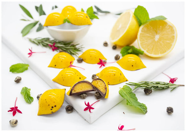Lemon Ginger bright yellow, lemon-shaped dark chocolate bonbons on a white table next to sliced lemon and green herbs