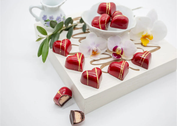 Red heart-shaped Lychee Bloom dark chocolate bonbons with creamy lychee white chocolate filling on a white table with white flowers