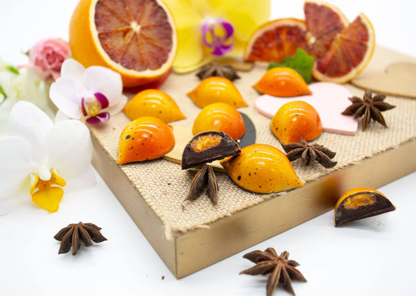Orange Star dark chocolate bonbon with ombre orange coloring and orange marmalade and dark chocolate ganache filling on white table with star anise and blood orange