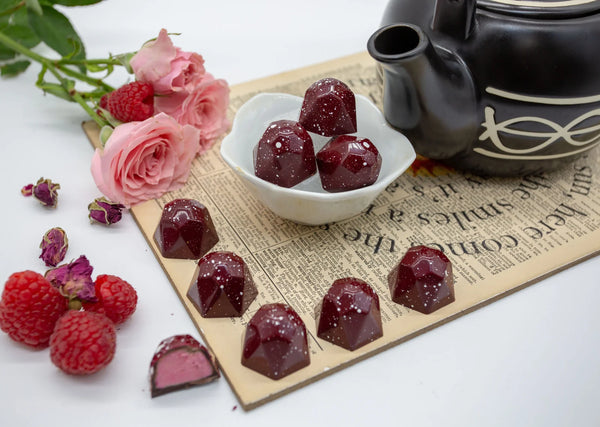Raspberry Rose deep red dark chocolate bonbons with pink raspberry filling on a white table with pink roses, a teapot, and raspberries