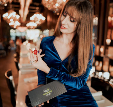 Beata in a velvet blue dress in front of a luxurious dining hall, holding a Sinless Treats gift box in one hand and a heart-shaped red Lychee Bloom chocolate in the other