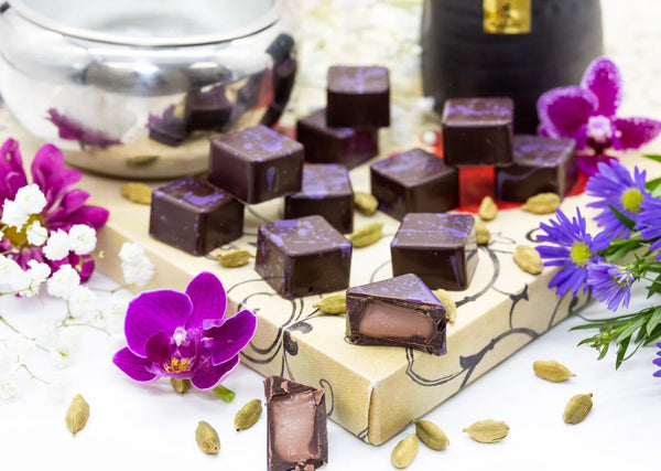 Cardamom Lava dark chocolate bonbons with purple splattered paint and creamy cardamom filling on a white table surrounded by purple and white flowers and cardamom pods