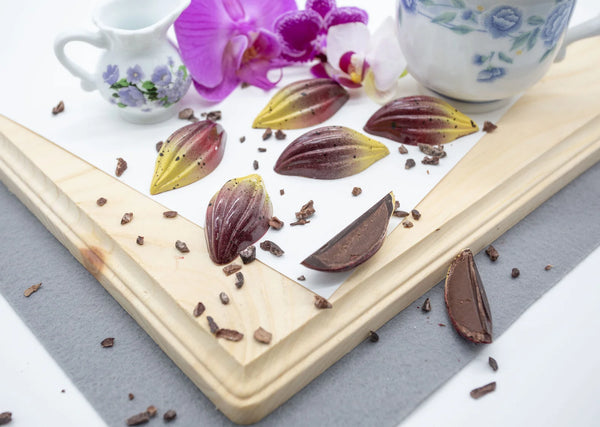 Dark chocolate bonbons shaped like cacao fruit with red and yellow coloring and creamy dark chocolate ganache on a white table with flowers