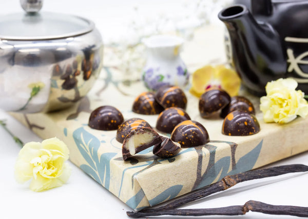 Dulce de Cream dark chocolate dome-shaped bonbons with orange splattered paint and milky vanilla filling on a white table next to white flowers, vanilla plant, an a teapot