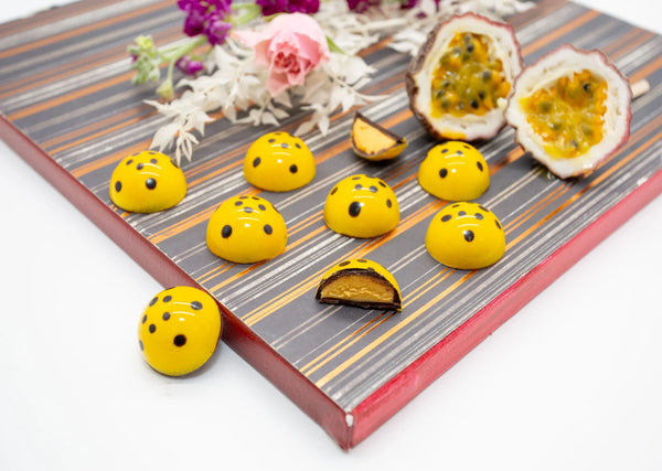 Yellow dome-shaped Maracuya dark chocolate bonbons on a white table with a cut-open passion fruit and flowers