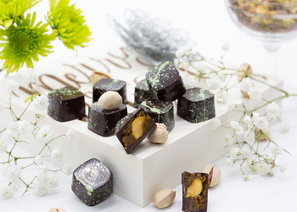 Pistachio Caramel dark chocolate bonbons with green splattered paint on a white table surrounded by white flowers and pistachios
