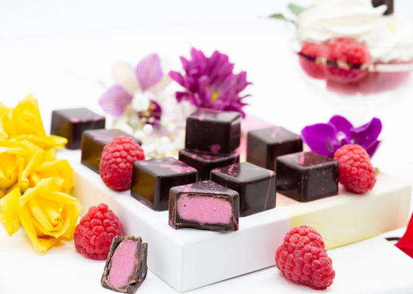 Roses and Raspberries dark chocolate bonbons with pink splattered paint and creamy pink filling on a white table surrounded by roses and raspberries