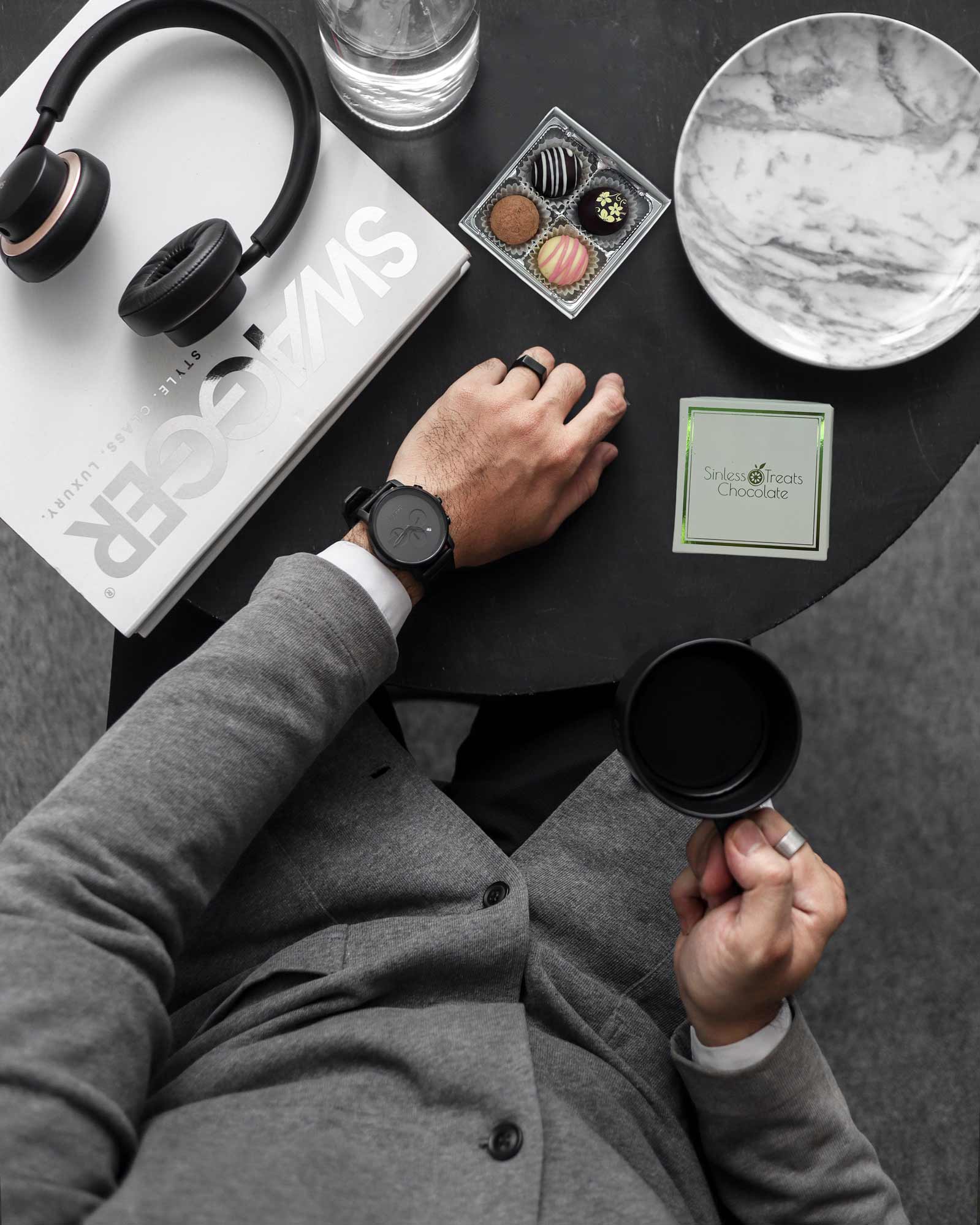 Overhead view of a business man holding a cup of coffee with his laptop, headphones, and 4-piece box of chocolate truffles at a small table